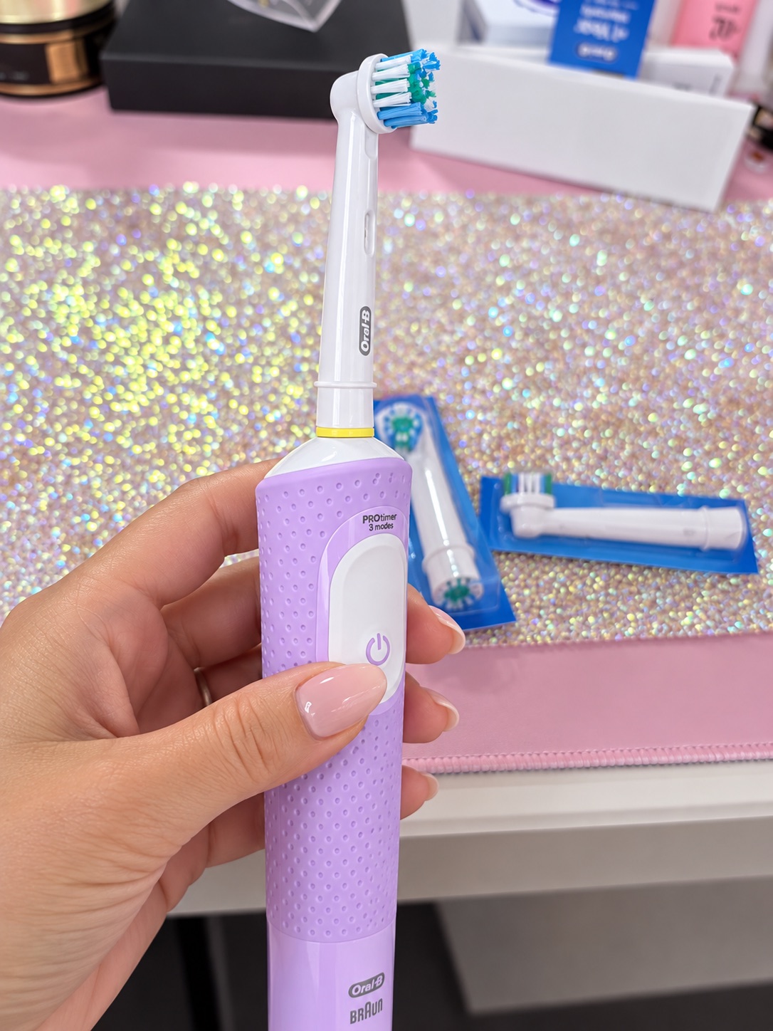 Hand holding the lilac Oral-B Vitality Pro showing the single power button and PRO timer label, with spare round brush heads in their packs behind